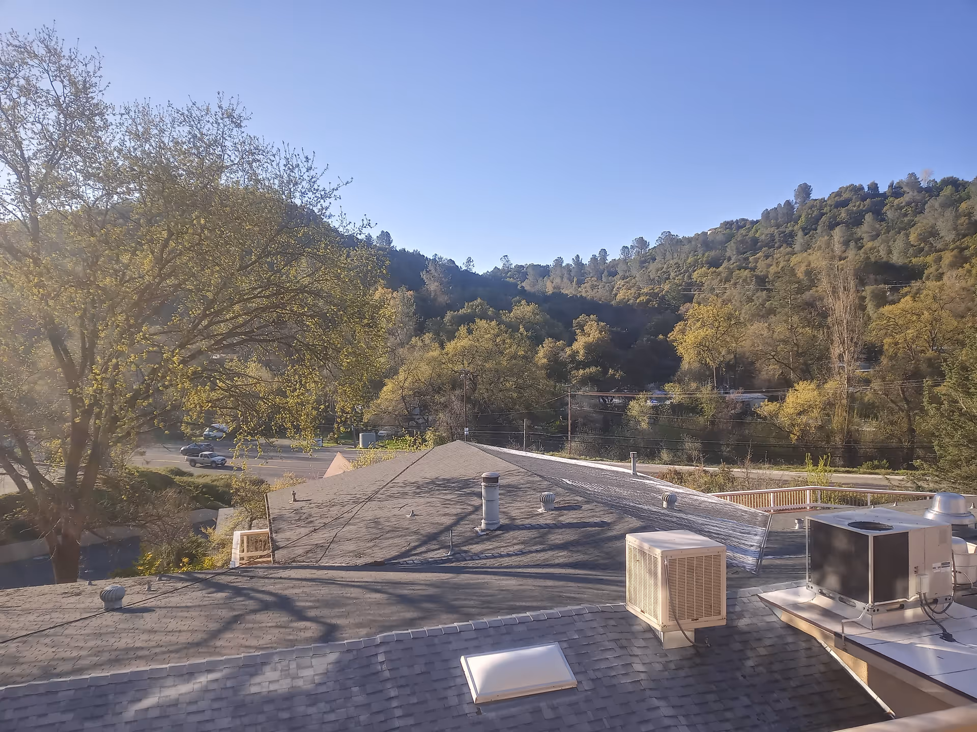 View of rooftops with HVAC units and vents, trees with green foliage, a road with cars, and forested hills under a clear blue sky.