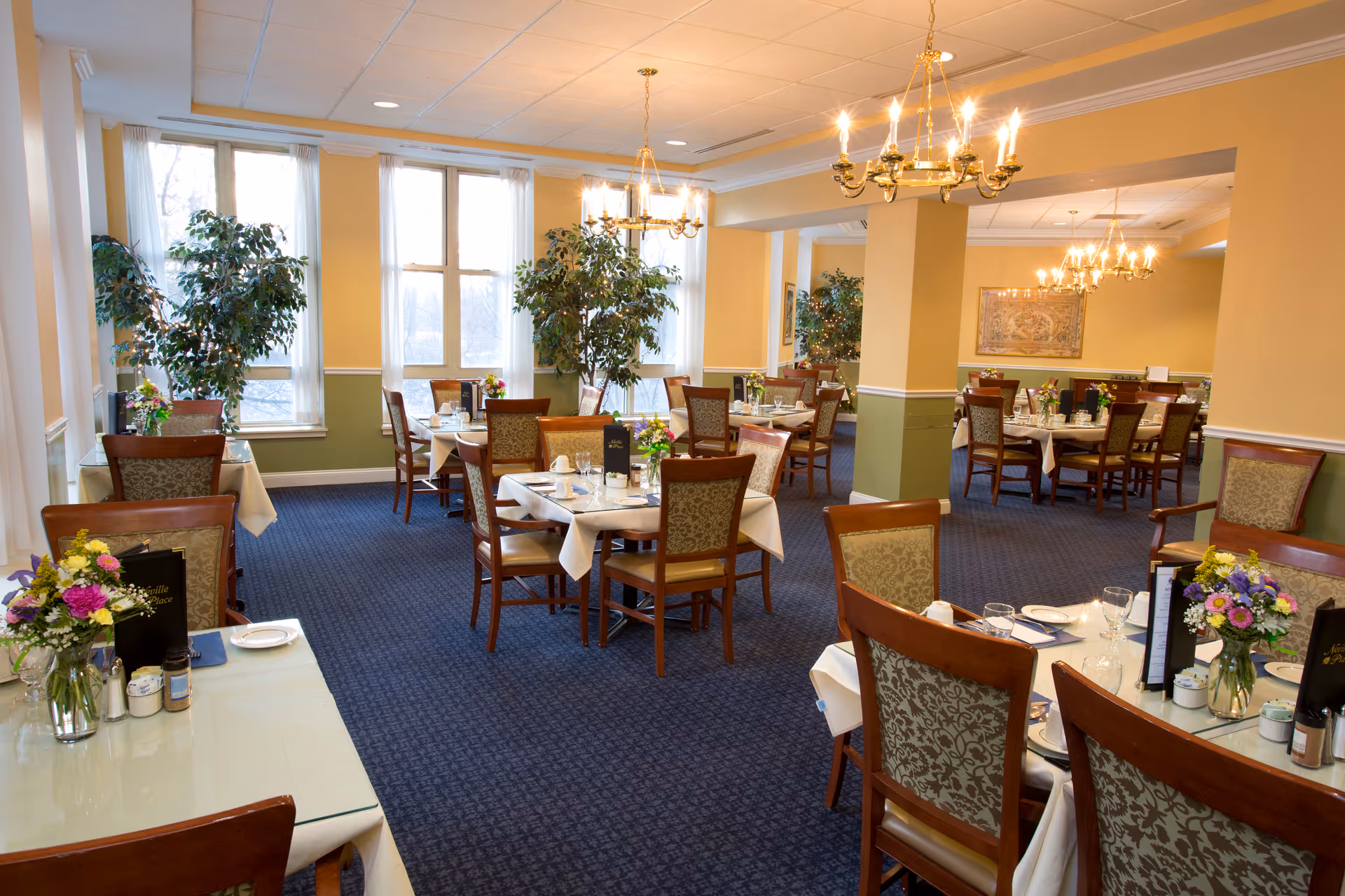 A spacious dining room with multiple tables set for meals, featuring white tablecloths, floral centerpieces, and wooden chairs with patterned upholstery. The room has large windows with sheer white curtains, blue carpeted floors, yellow walls with green wainscoting, and elegant chandeliers hanging from the ceiling. Several potted plants are placed near the windows and columns.