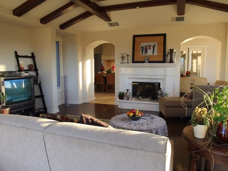 A cozy living room with a beige sofa facing a white fireplace. There is a round ottoman with a bowl of fruit on it in the center. To the right, there is a side table with several potted plants. The room has wooden beams on the ceiling and an archway leading to a dining area with chairs and a table. A TV on a stand is positioned to the left side of the room.
