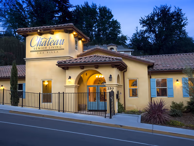 Exterior view of Chateau Senior Living The Villa building at dusk, featuring a yellow stucco facade with blue shutters, a tiled roof, and an arched entrance with blue double doors. The building is surrounded by a black metal fence and landscaping with trees and shrubs.