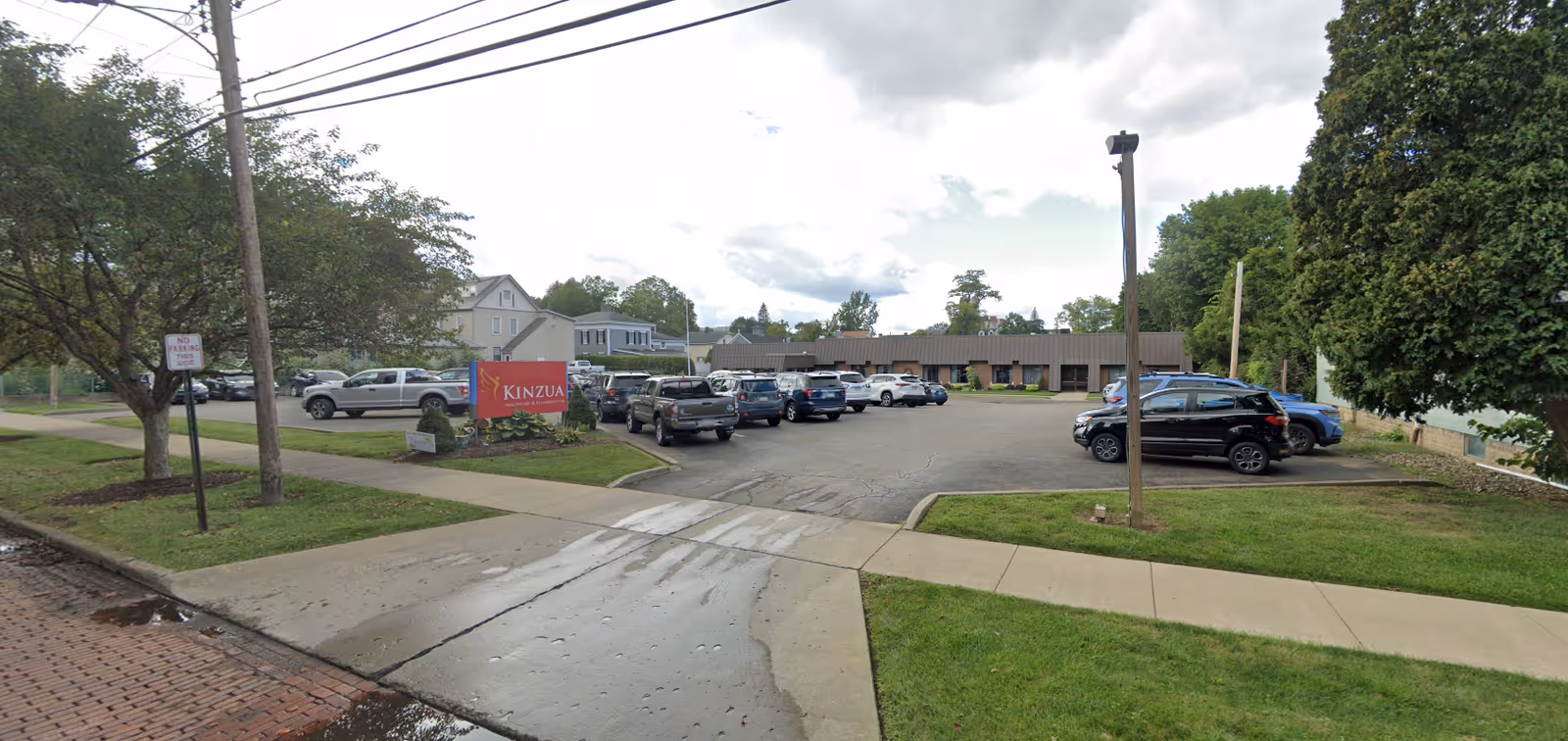 Exterior view of Kinzua Nursing and Rehabilitation facility showing a parking lot with several parked cars, a red sign with the facility name, trees, and a sidewalk under a partly cloudy sky.