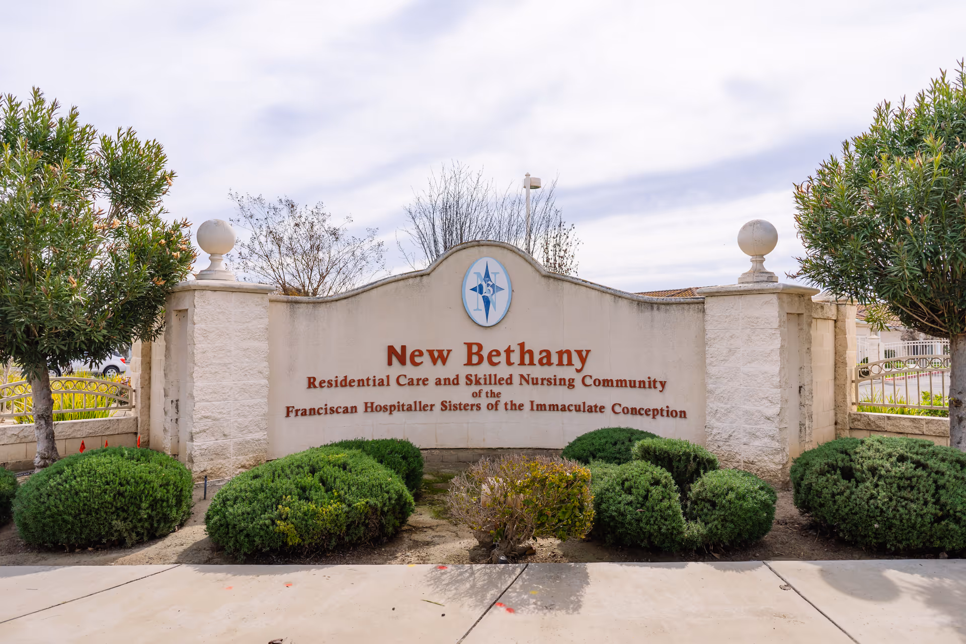 Entrance sign for New Bethany Residential Care and Skilled Nursing Community, surrounded by neatly trimmed bushes and trees, with a cloudy sky in the background.