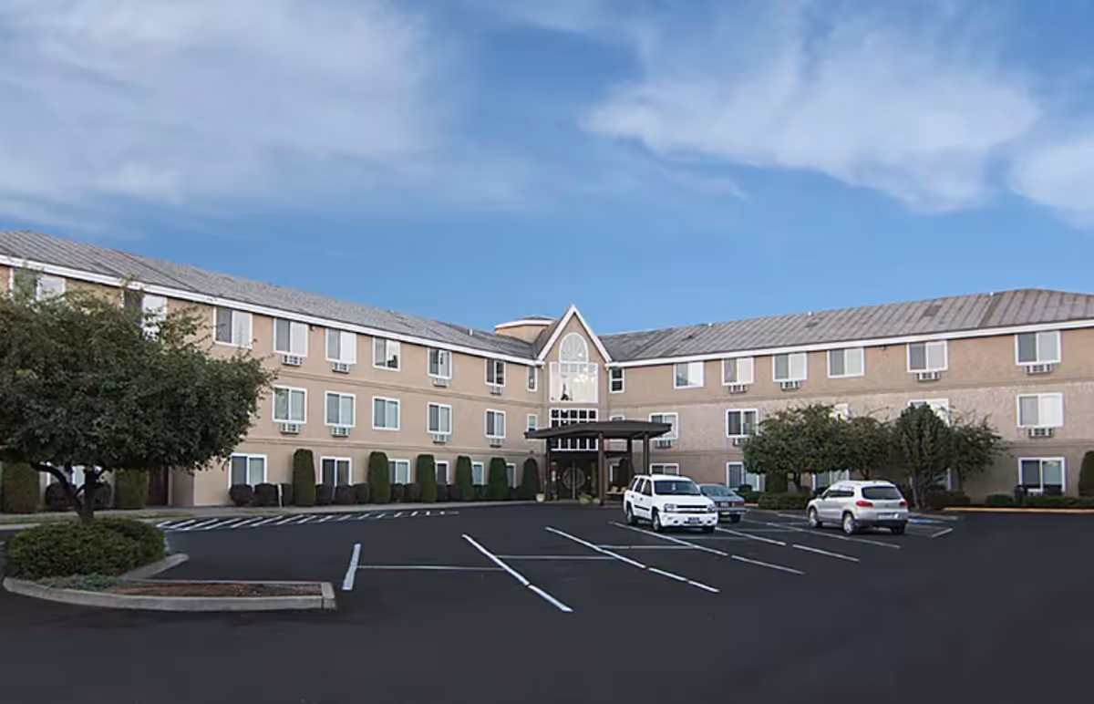 Exterior view of a three-story senior living facility building with multiple windows, a covered entrance, a parking lot with a few cars, and landscaped greenery including trees and bushes under a partly cloudy blue sky.