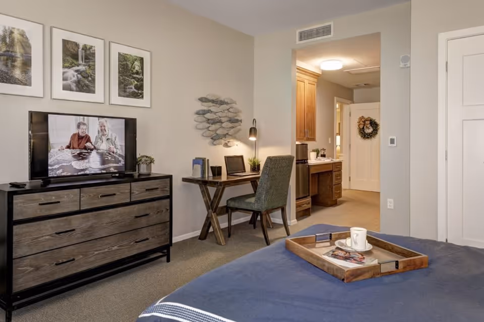 A cozy bedroom in a senior living facility featuring a bed with a wooden tray holding a cup and a magazine. Across from the bed is a wooden dresser with a flat-screen TV displaying two elderly people working on a puzzle. Above the TV are three framed nature photographs. To the right of the dresser is a small wooden desk with a green upholstered chair, a desk lamp, and some books. In the background, there is a hallway leading to a bathroom with wooden cabinets and a door decorated with a wreath.