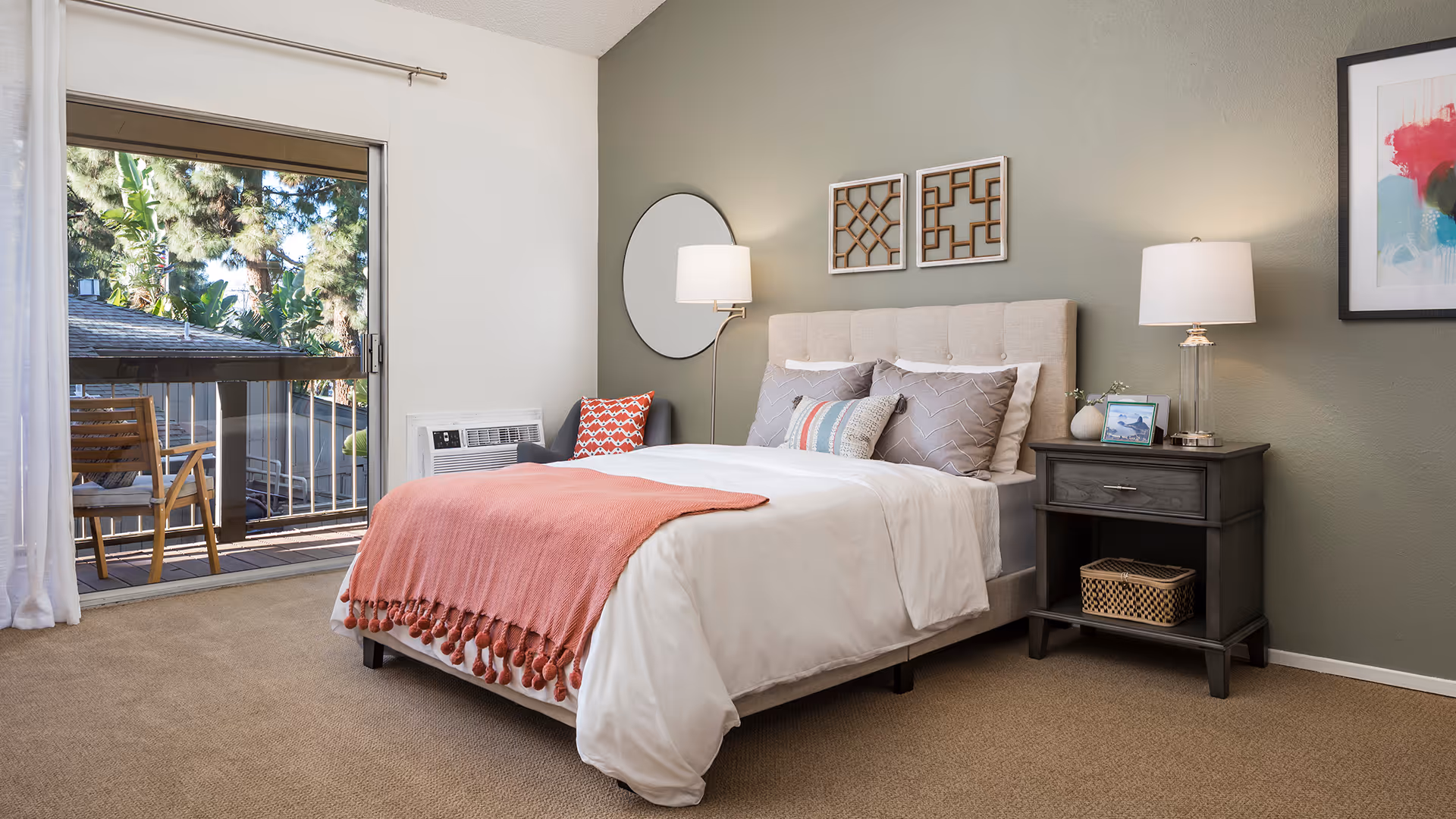 A cozy bedroom with a neatly made bed featuring white bedding and a coral throw blanket. There is a gray armchair with a red patterned pillow next to a sliding glass door that opens to a balcony with wooden chairs. The room has a green accent wall with two decorative wall hangings, a round mirror, and a nightstand with a lamp, a framed photo, and a small basket underneath.