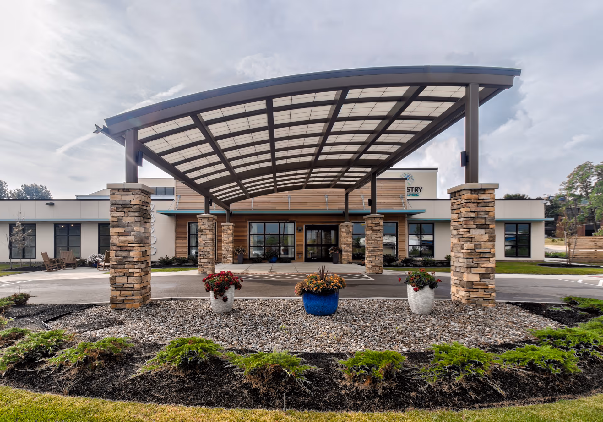 Front exterior view of a modern senior living facility with a large covered entrance supported by stone pillars. There are three flower pots with colorful flowers placed on a bed of rocks in front of the entrance. The building has large windows and a combination of wood and light-colored siding.