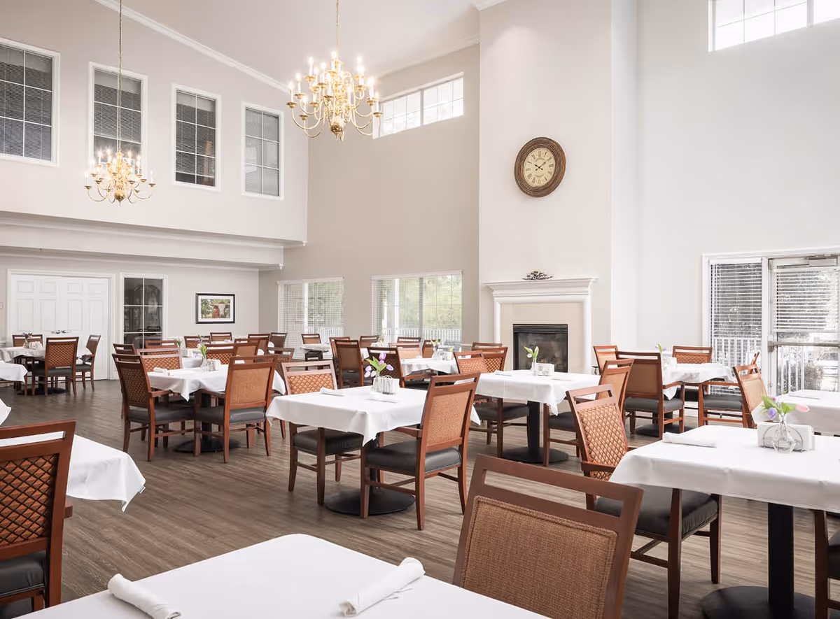 A bright and spacious dining room with multiple tables covered in white tablecloths, each set with napkins and small flower vases. The room features high ceilings with large windows, two chandeliers, a fireplace with a clock above it, and wooden chairs with cushioned seats.
