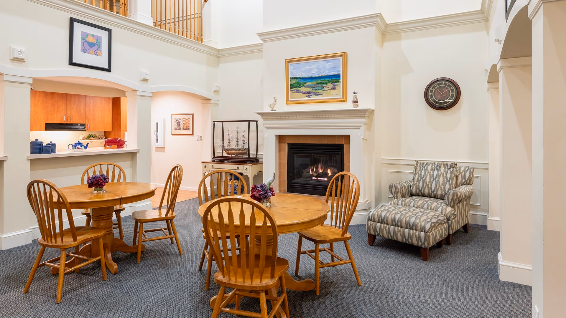 A cozy common area in a senior living facility featuring two round wooden tables each surrounded by four wooden chairs. There is a fireplace with a painting above it, a decorative ship model on a cabinet, and a patterned armchair with an ottoman. The walls are painted white, and the carpet is gray. The kitchen area is visible through a pass-through window.