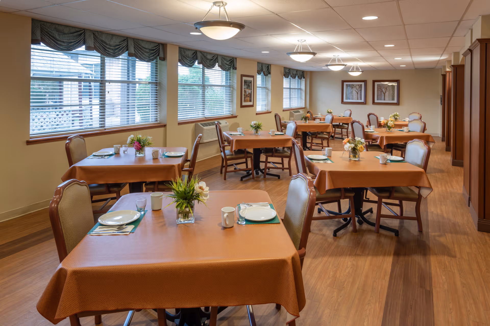 A dining room with multiple tables covered in orange tablecloths, each set with plates, cups, and small flower arrangements. The room has wooden flooring, large windows with blinds and valances, and framed artwork on the walls. Ceiling lights provide warm illumination.
