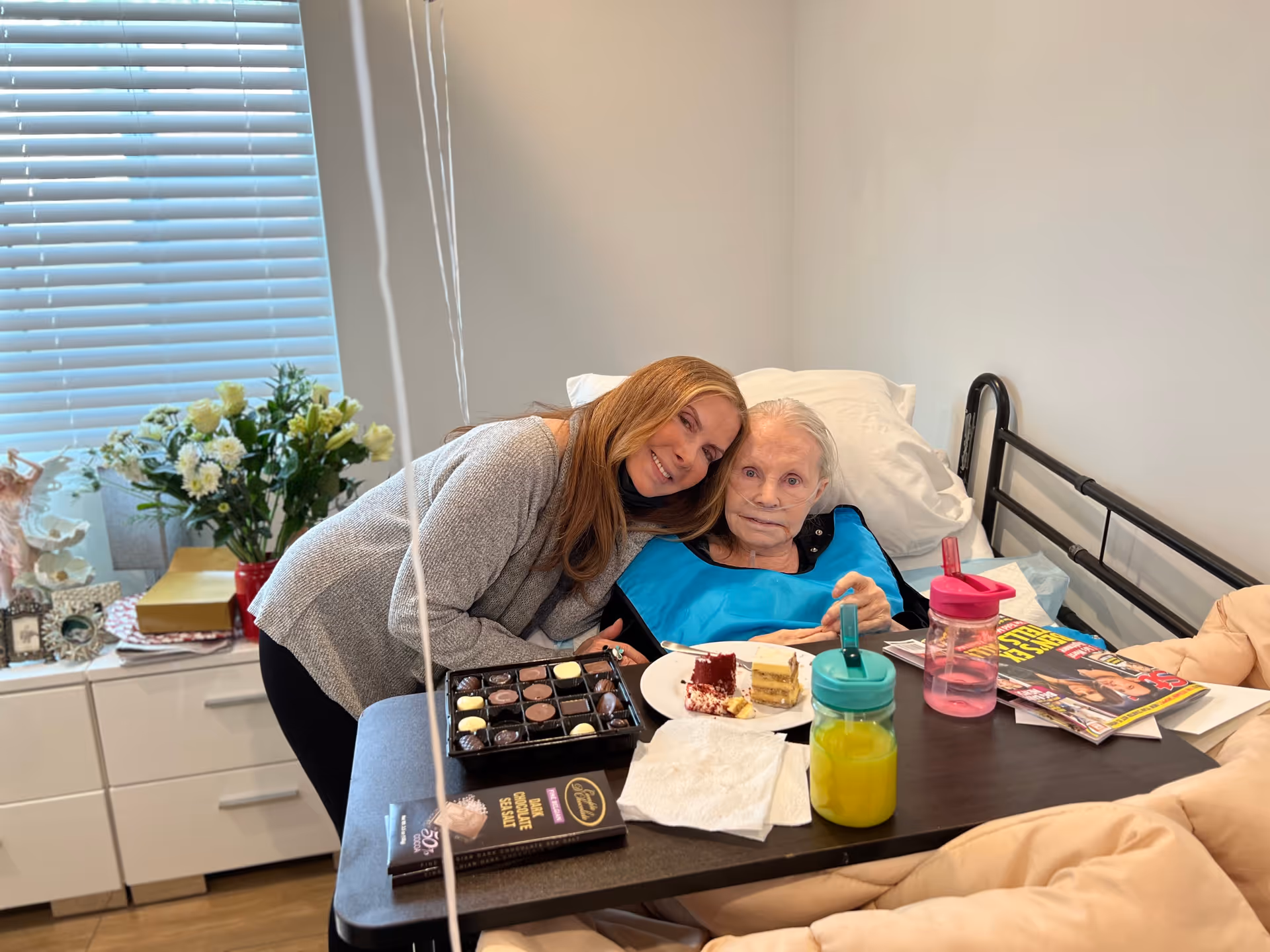 An elderly woman lying in a hospital-style bed with a blue bib, accompanied by a younger woman leaning close to her and smiling. On the bedside table, there are plates with cake, a box of chocolates, two colorful drink containers, magazines, and napkins. In the background, there is a window with blinds and a cabinet with flowers and framed photos.