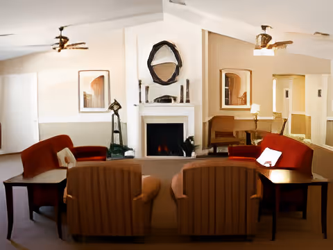 A cozy living room with two beige armchairs and two red sofas arranged around a white fireplace. The fireplace has a round mirror and decorative items on the mantel. There are ceiling fans, framed artwork on the walls, and side tables next to the seating.