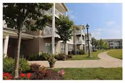 Exterior view of a multi-story senior living facility with balconies, a paved walkway, landscaped greenery, trees, and a clear blue sky.