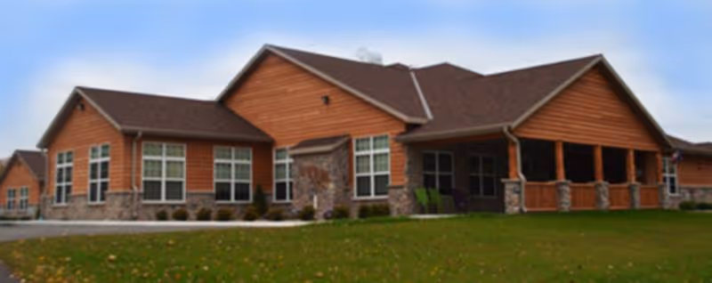 Exterior view of a single-story senior living facility building with brown wooden siding and stone accents. The building has multiple large windows and a covered porch area with seating. The surrounding area includes a green lawn and a cloudy sky.