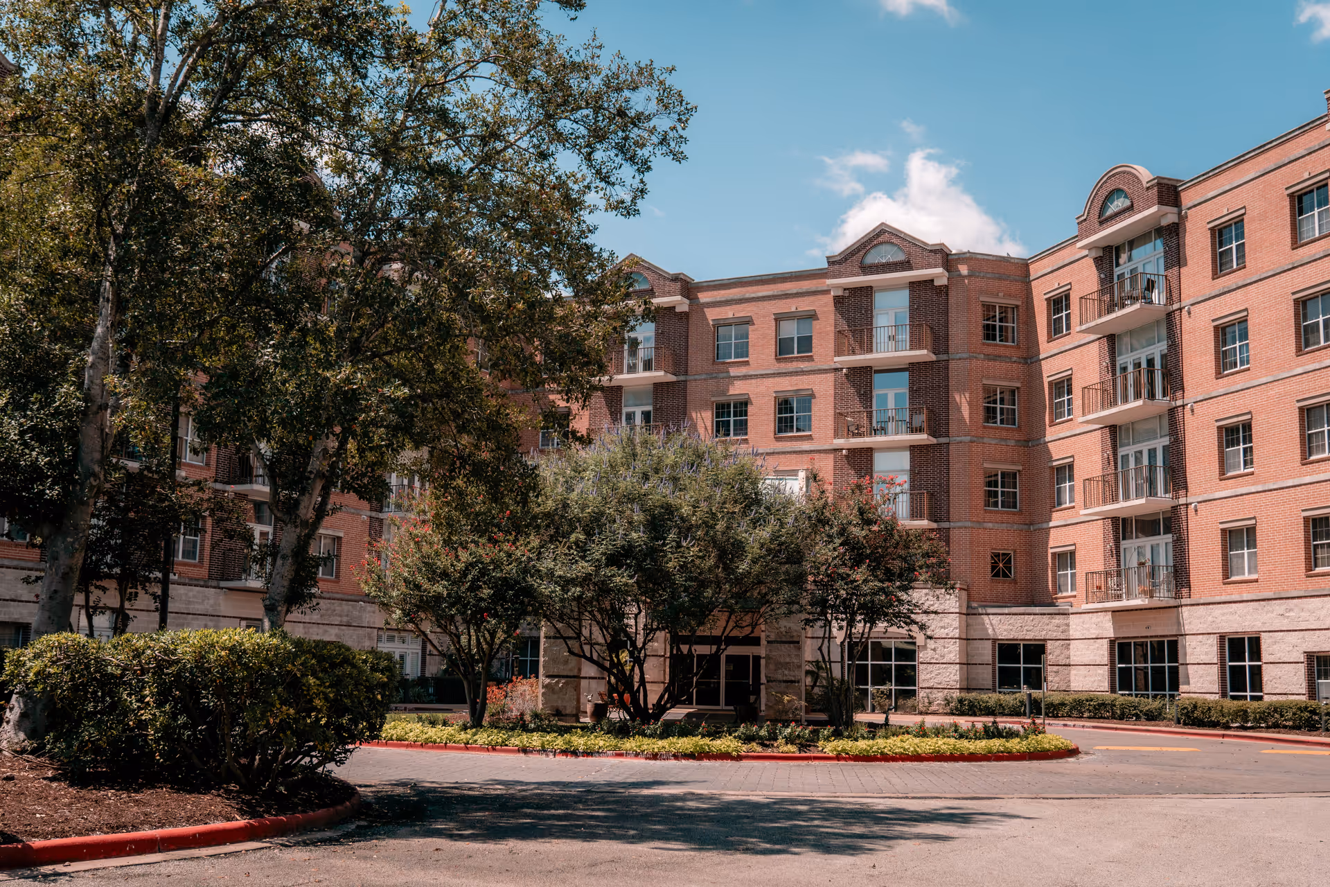 Exterior view of a multi-story brick building with several windows and balconies, surrounded by trees and landscaped greenery under a blue sky with some clouds.