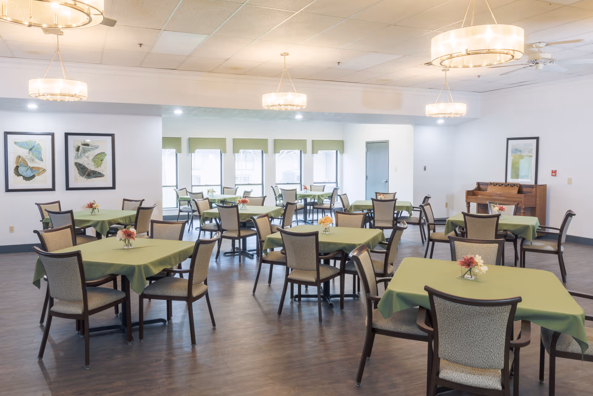 A bright dining room with multiple square tables covered with green tablecloths, each table surrounded by four chairs. Small floral centerpieces are placed on each table. The room has large windows with green valances, framed butterfly artwork on one wall, a piano against another wall, and modern circular ceiling lights.