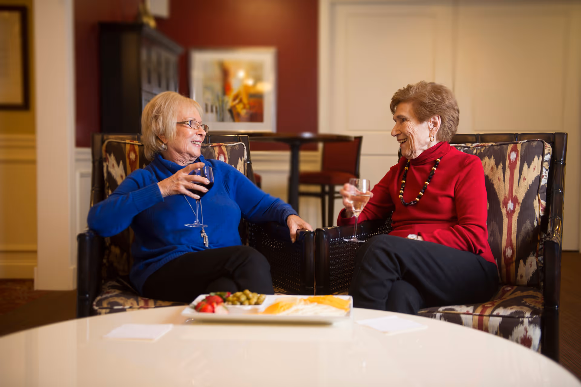 Two elderly women sitting in patterned armchairs in a cozy indoor setting, smiling and holding wine glasses while conversing. A white table in front of them holds a plate with cheese, olives, and strawberries. The background features warm-colored walls, framed artwork, and additional seating.
