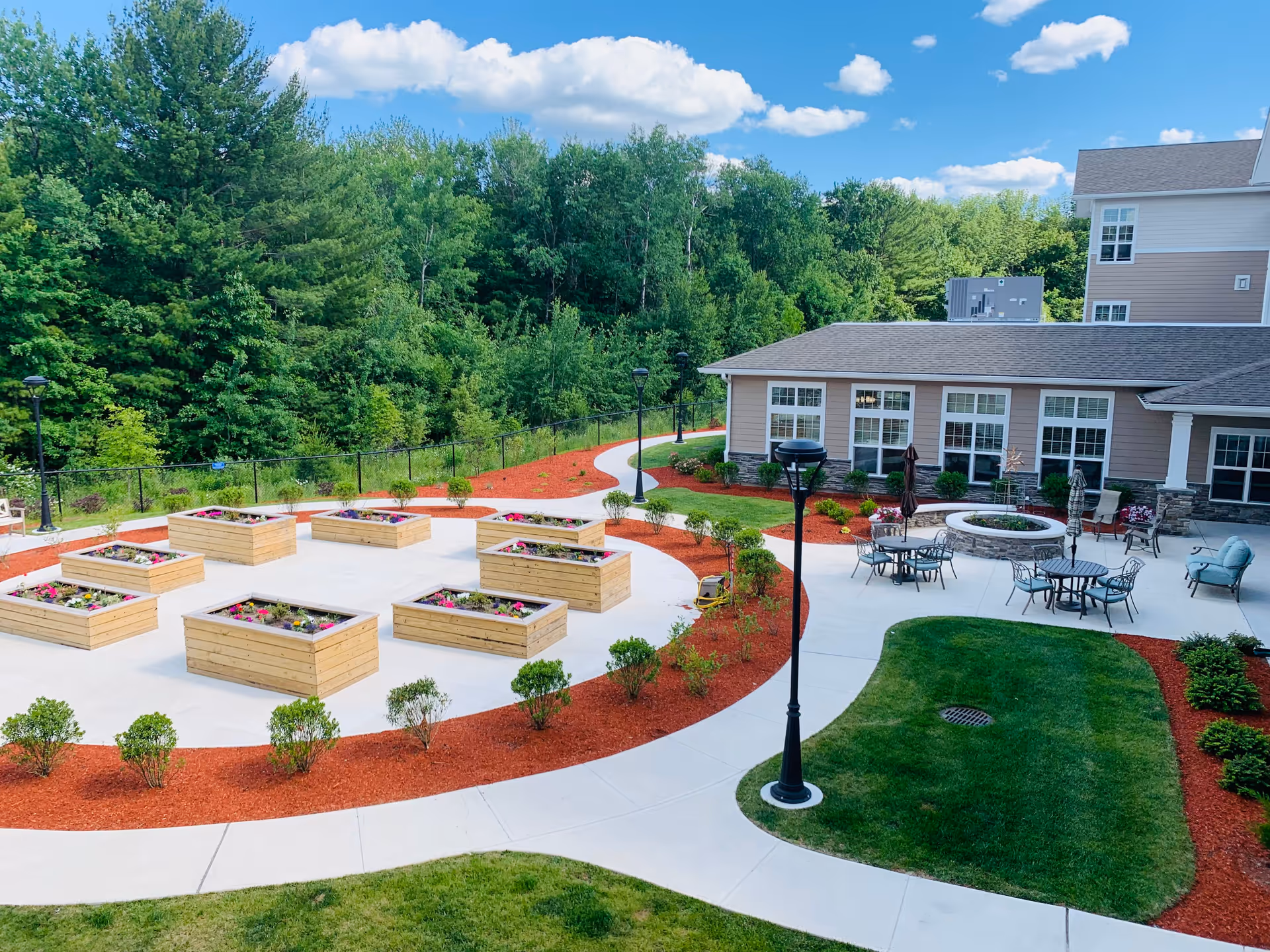 Outdoor patio area at Kennedy Meadows Gracious Retirement Living featuring raised wooden garden beds with flowers, a circular fire pit surrounded by tables and chairs, well-maintained green lawns, landscaped bushes, and a backdrop of dense green trees under a blue sky with scattered clouds.