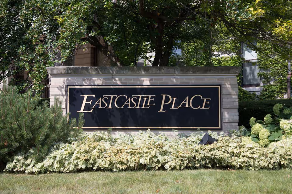 A large stone sign with a black background and gold lettering that reads 'Eastcastle Place,' surrounded by green bushes and trees in a landscaped outdoor area.
