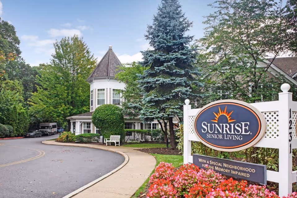 Exterior view of Sunrise Senior Living facility with a curved driveway, landscaped greenery, and a white sign displaying the Sunrise Senior Living logo and a message about a special neighborhood for the memory impaired.
