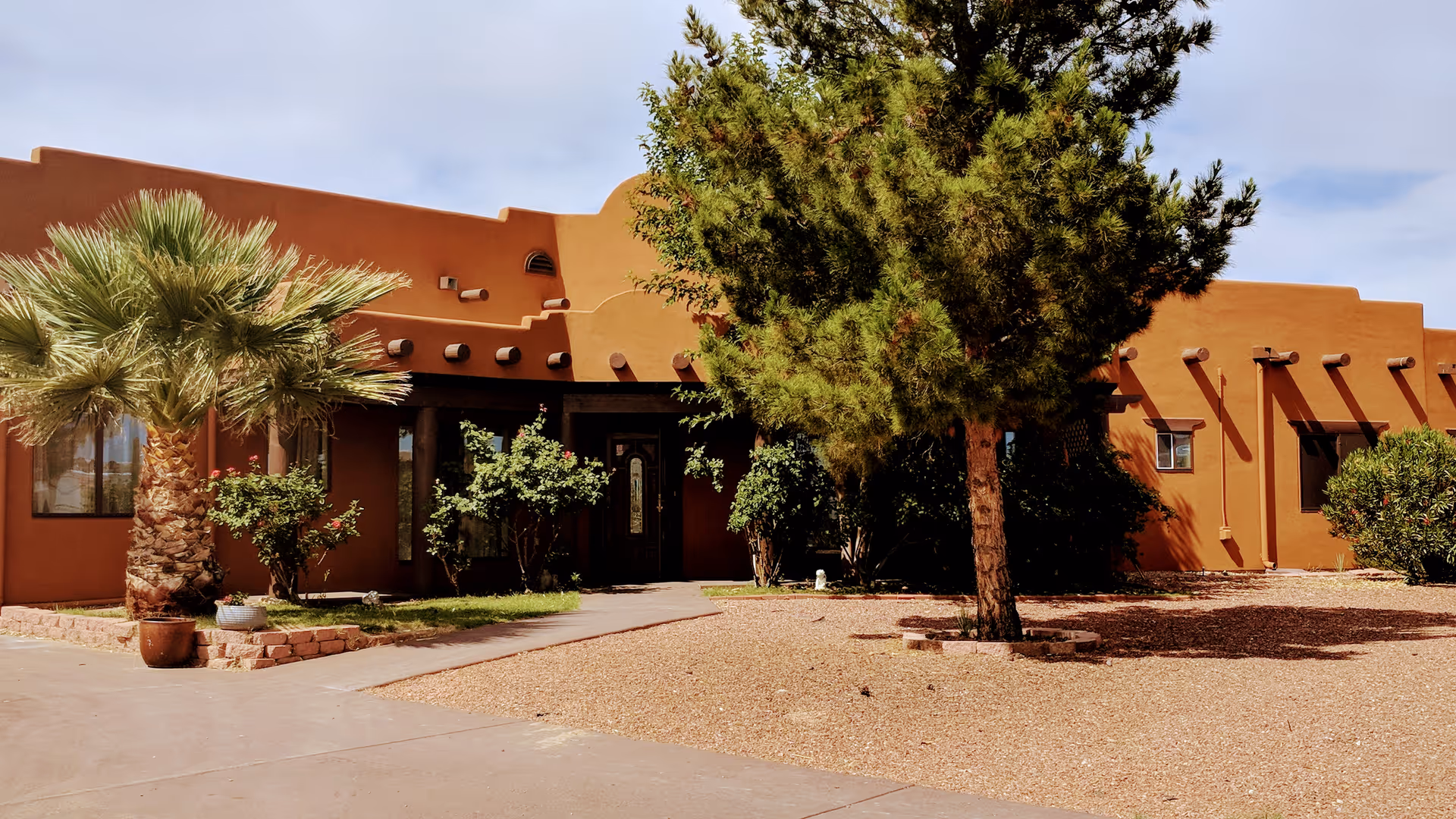 Exterior view of Casa De Angeles Assisted Living Inc, showing a southwestern-style building with brown stucco walls, a front entrance with glass doors, and surrounding landscaping including palm trees, bushes, and a large pine tree. The ground is covered with gravel and there is a paved walkway leading to the entrance.