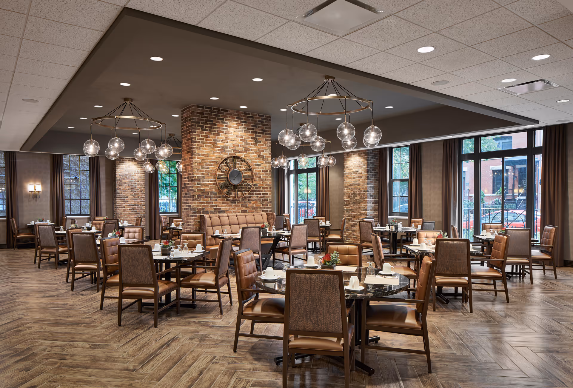 A spacious and elegant dining room in a senior living facility featuring multiple round and square tables with brown cushioned chairs. The room has large windows with brown curtains, exposed brick pillars, modern spherical chandeliers, and a large decorative wall clock. The tables are set with white cups, plates, and small flower arrangements.
