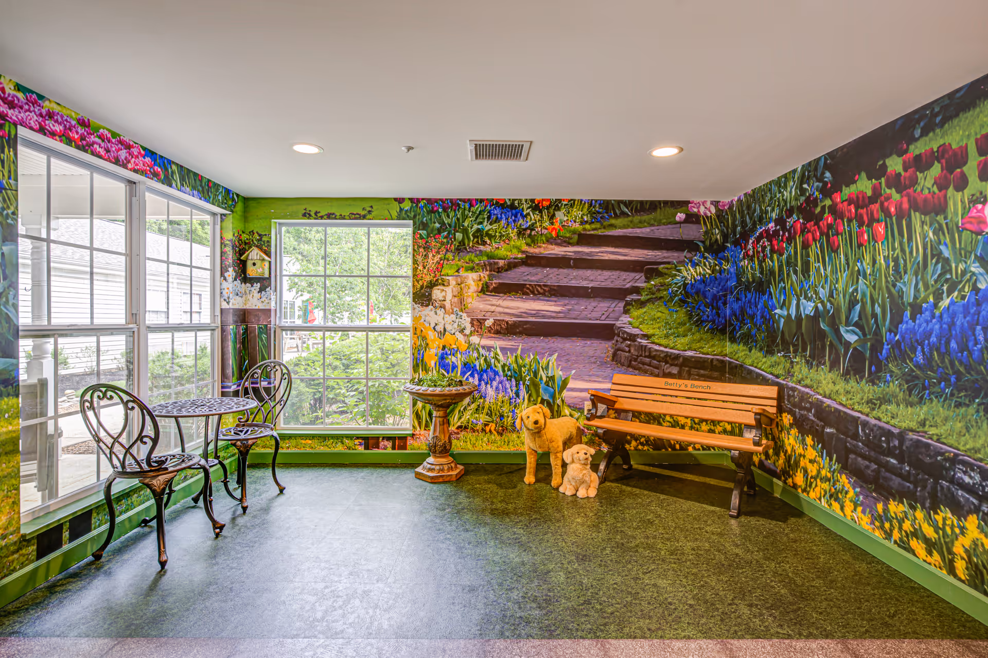 A bright indoor room with large windows and walls covered in a mural of a colorful garden with flowers and stone steps. The room contains a small metal table with two matching chairs, a wooden bench labeled 'Betty's Bench', a decorative birdbath, and two stuffed dog figures on the floor.