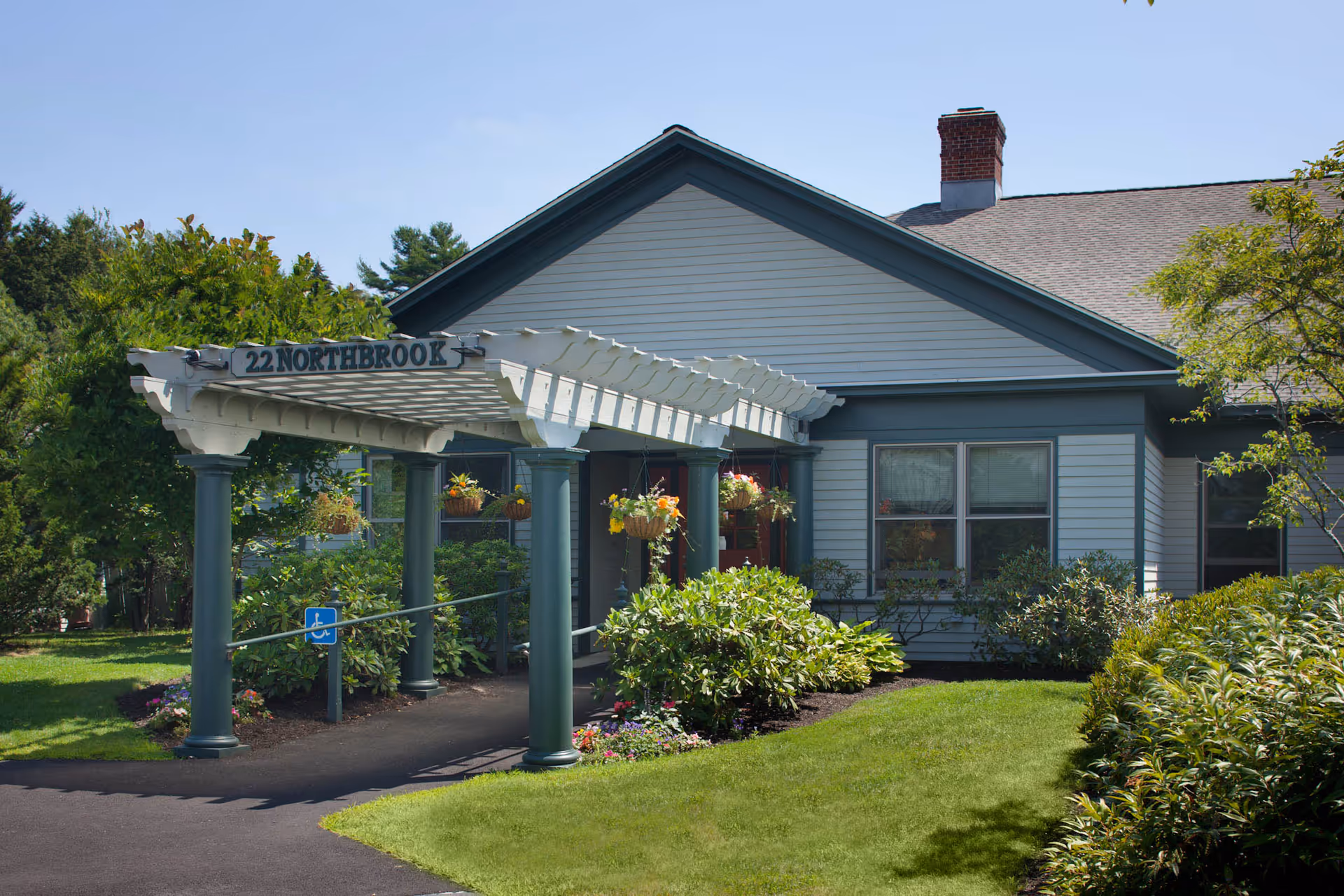 Exterior view of a single-story building with light gray siding and a dark gray roof. A white pergola supported by green columns extends over a paved walkway leading to the entrance. Hanging flower baskets and well-maintained bushes and lawn surround the building. A sign on the pergola reads '22 NORTHBROOK'.