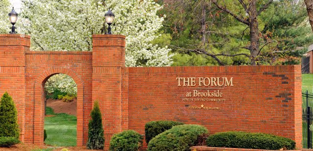 Brick entrance wall with an archway and two pillars topped with lanterns, surrounded by green bushes and trees with white blossoms. The wall has a sign that reads 'THE FORUM at Brookside Senior Living Community'.