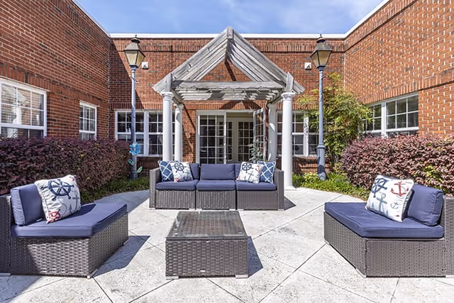 Outdoor patio area at Brookdale Wilmington with wicker seating furniture including a sofa and two chairs with navy blue cushions and nautical-themed pillows. The patio is surrounded by red brick walls, two lampposts, and greenery. A wooden pergola structure is positioned above the sofa near the entrance doors.