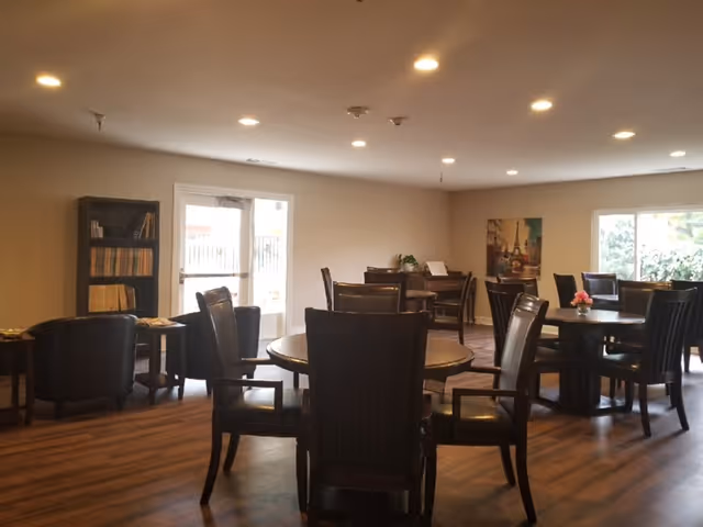 A well-lit room with multiple round tables surrounded by dark wooden chairs with armrests. There is a bookshelf filled with books on the left side near a glass door leading outside. The room has wooden flooring, beige walls, and ceiling lights. A window on the right side shows greenery outside, and a framed picture hangs on the wall.