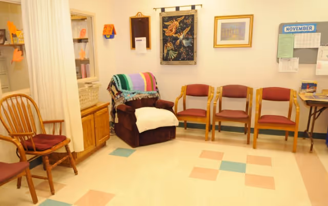 A small waiting or sitting area with a brown recliner covered by a colorful blanket and a white cloth, three wooden chairs with red cushions, a wooden chair with a red cushion, a bulletin board with a November calendar, and framed artwork on the walls.