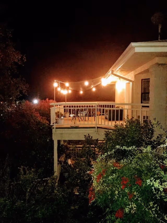 A nighttime view of a balcony or patio area illuminated by string lights. The balcony has a white railing and contains a few chairs and potted plants. Surrounding the balcony are dense bushes and trees with some red flowers visible.