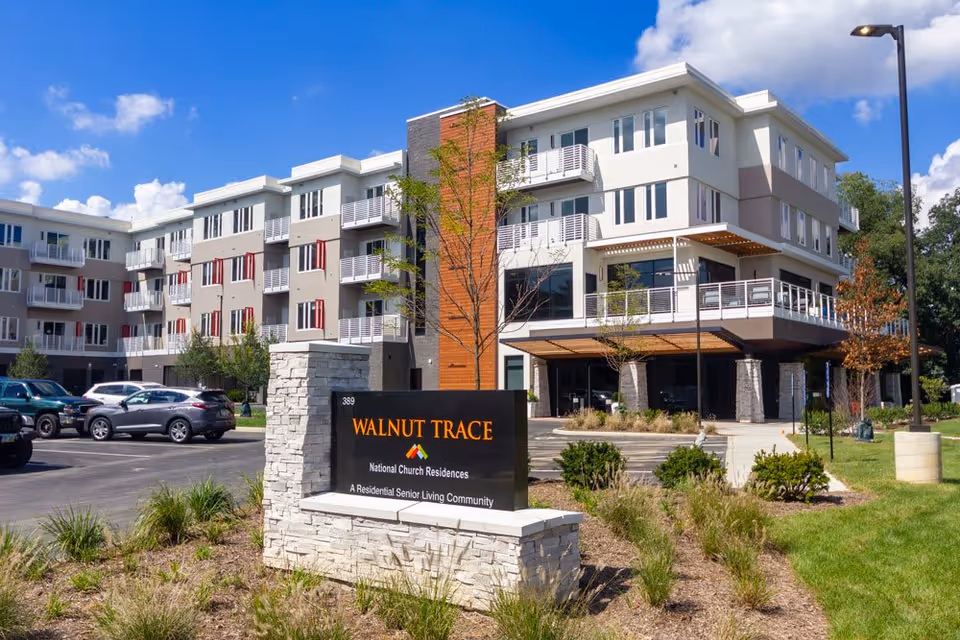 Exterior view of Walnut Trace, a modern multi-story senior living community building with balconies, surrounded by landscaped greenery and a parking lot with cars. A stone sign in the foreground reads 'Walnut Trace, National Church Residences, A Residential Senior Living Community.'