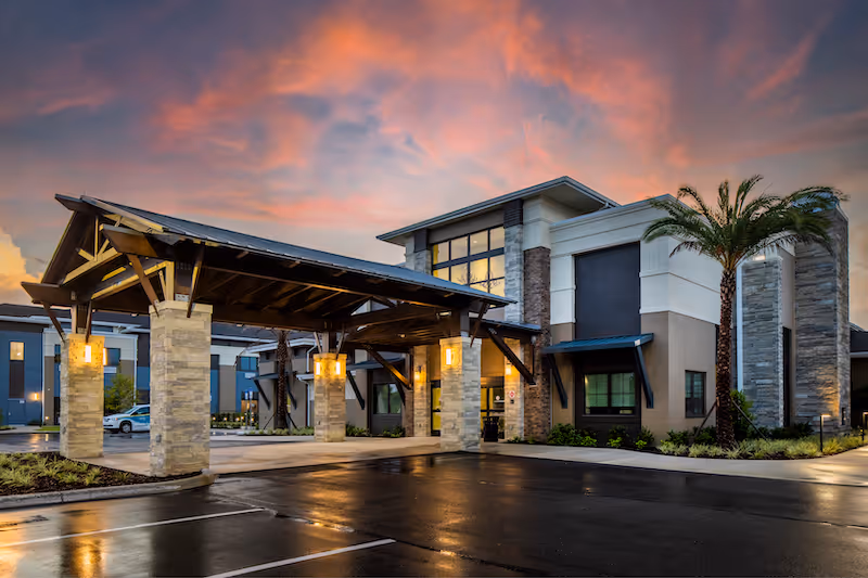 Exterior view of a modern senior living facility building at sunset with a covered entrance supported by stone pillars, palm trees, and a wet parking lot reflecting the lights.