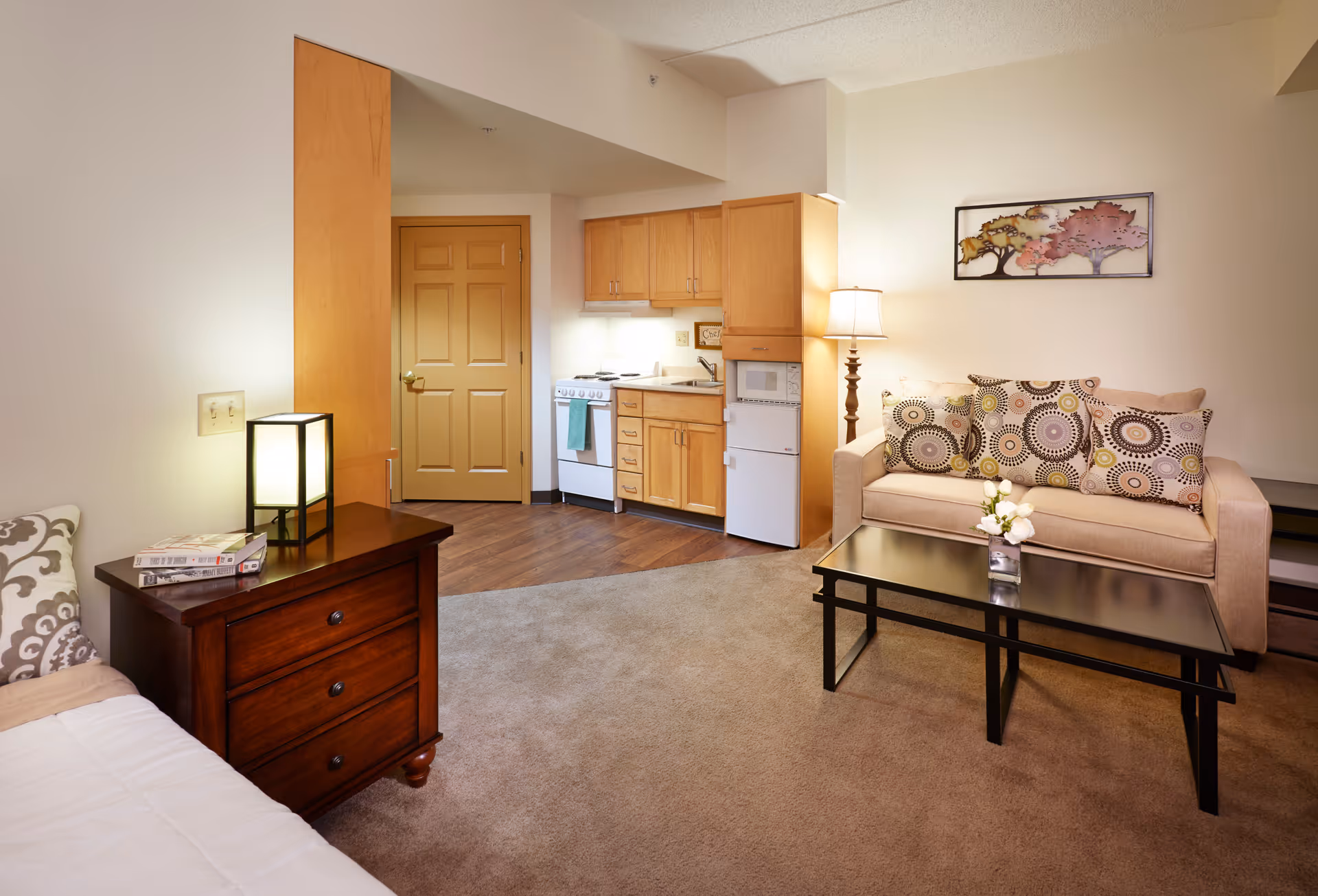 A cozy living area in a senior living facility featuring a beige sofa with patterned cushions, a black coffee table with a small flower vase, a wooden side table with a lamp and books, and a compact kitchen area with wooden cabinets, a stove, microwave, and refrigerator. The room has carpeted and wooden flooring, a beige door, and a framed artwork of trees on the wall.