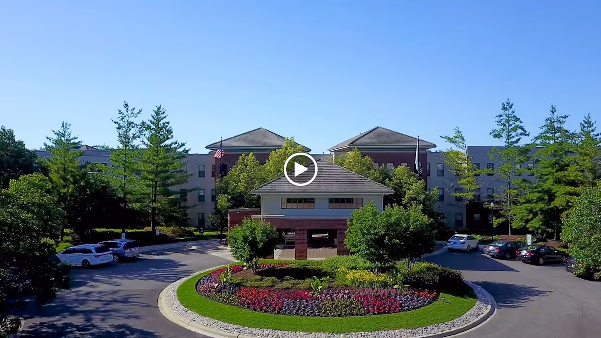Front exterior view of the Wyndemere senior living facility with a circular driveway, landscaped garden with colorful flowers, trees, and several parked cars under a clear blue sky.