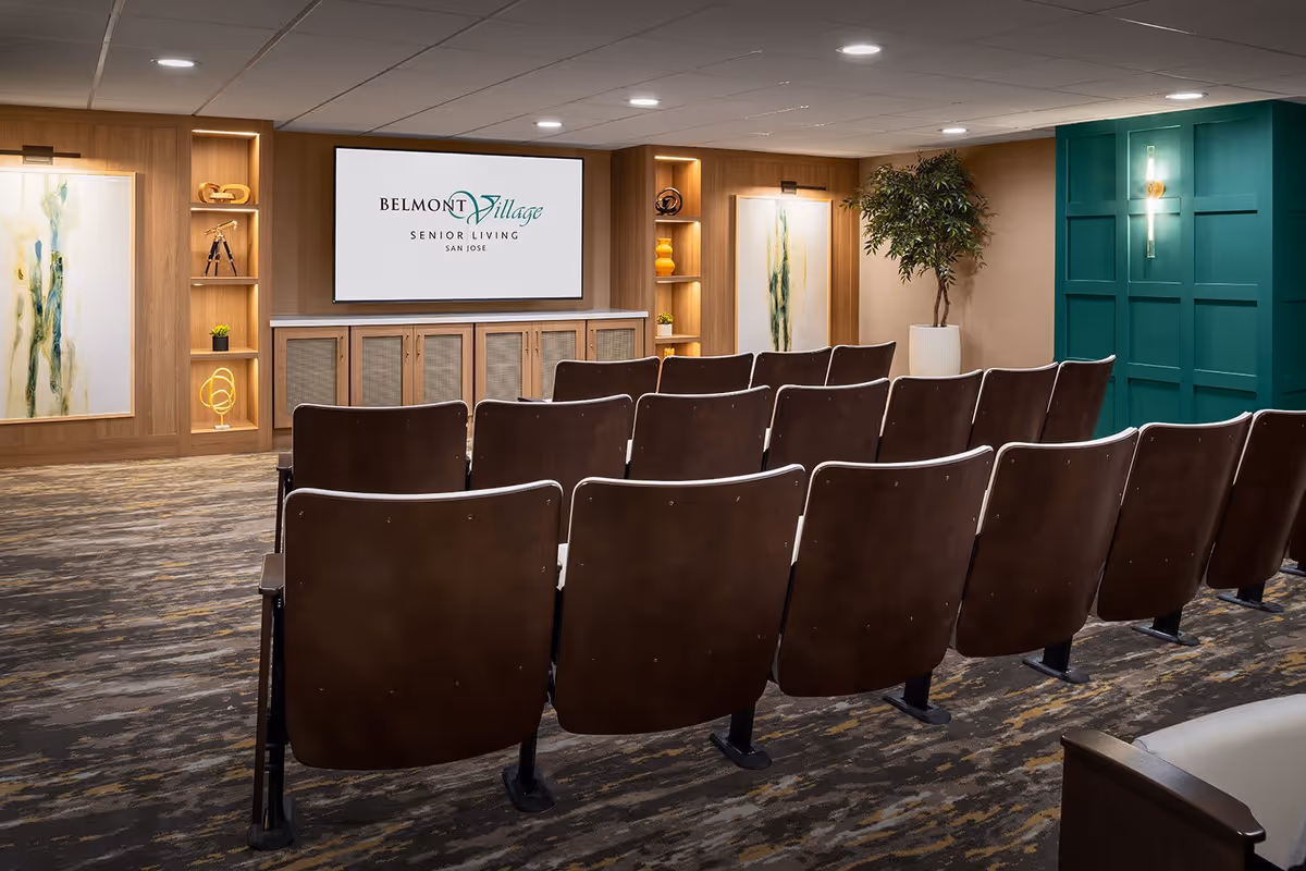 A small theater or presentation room with rows of brown chairs facing a large screen displaying the Belmont Village Senior Living San Jose logo. The room features wood paneling, built-in shelves with decorative items, framed artwork, a potted plant, and a teal accent wall with a sconce light.