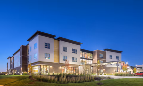 Exterior view of a modern three-story senior living facility building at dusk with well-lit windows, landscaped greenery, and a clear blue sky.