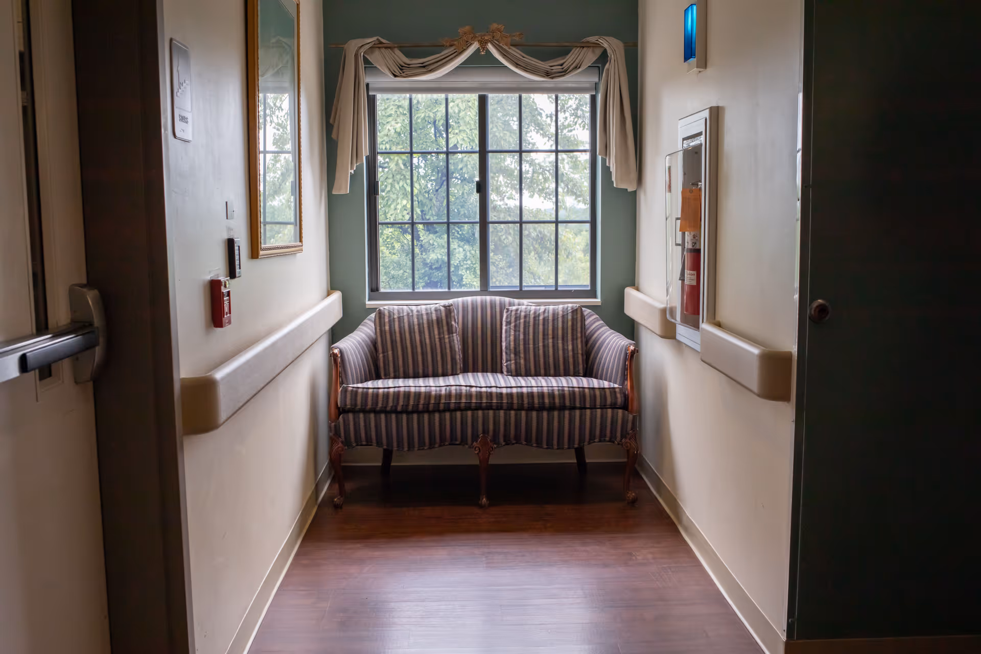 A small sitting area in a hallway with a striped loveseat positioned in front of a large window with a decorative curtain. The walls are light-colored with handrails on both sides, and there is a fire extinguisher cabinet mounted on the right wall.