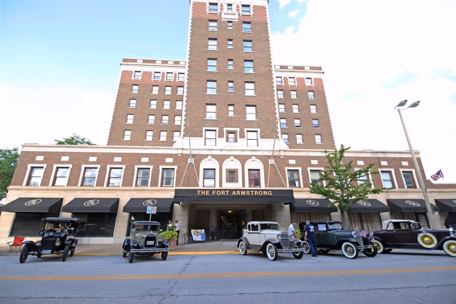 Front exterior view of The Fort Armstrong building, a multi-story brick structure with vintage cars parked in front and an American flag on the right side.