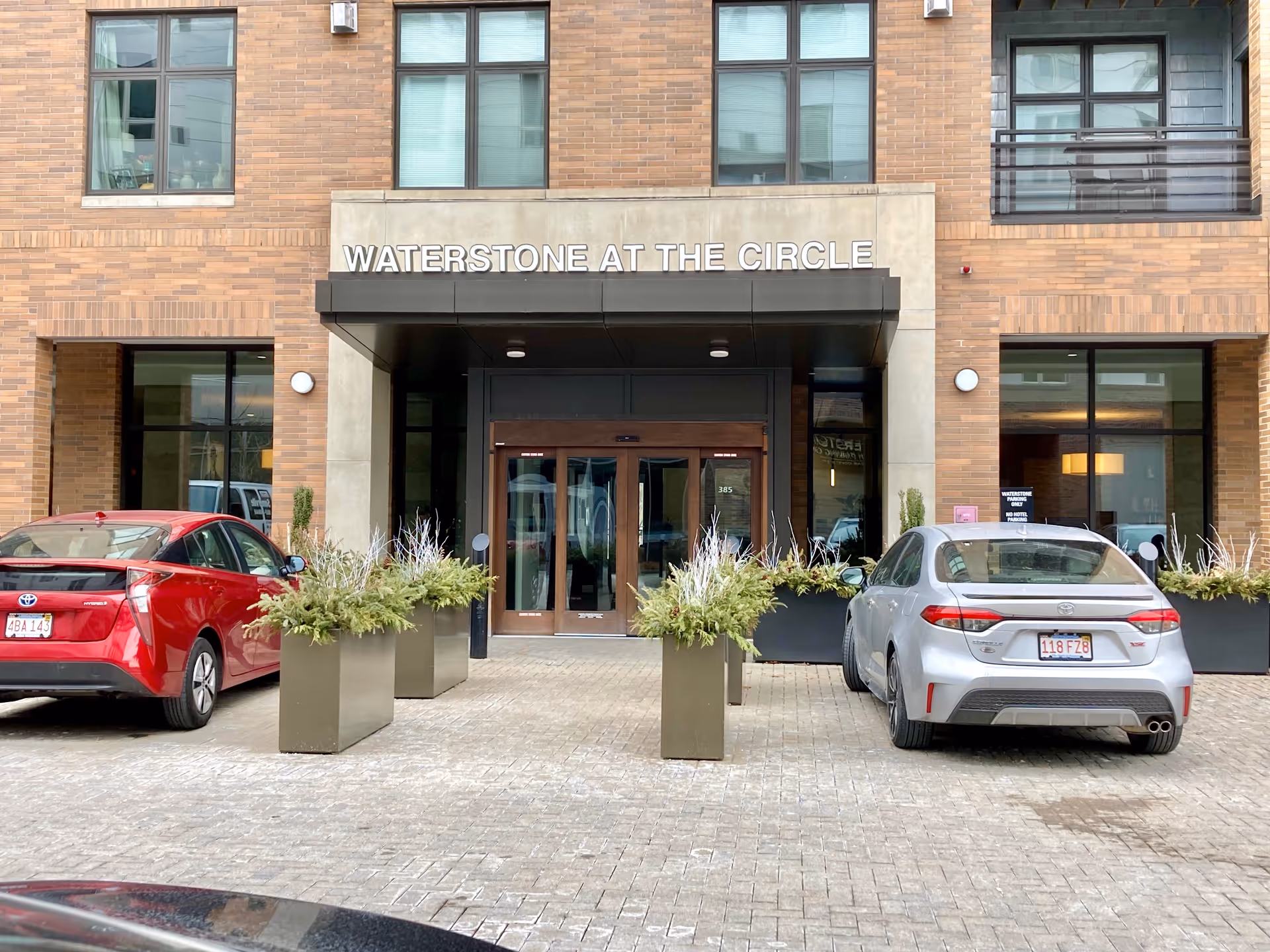 Entrance of Waterstone at the Circle facility showing a modern building facade with large windows, a covered doorway with glass double doors, and two cars parked in front. There are tall planters with greenery lining the walkway to the entrance.