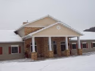 Front exterior view of a single-story assisted living facility building with a covered entrance supported by stone pillars. The ground and roof are covered with snow, and the building has beige siding with red shutters on the windows.
