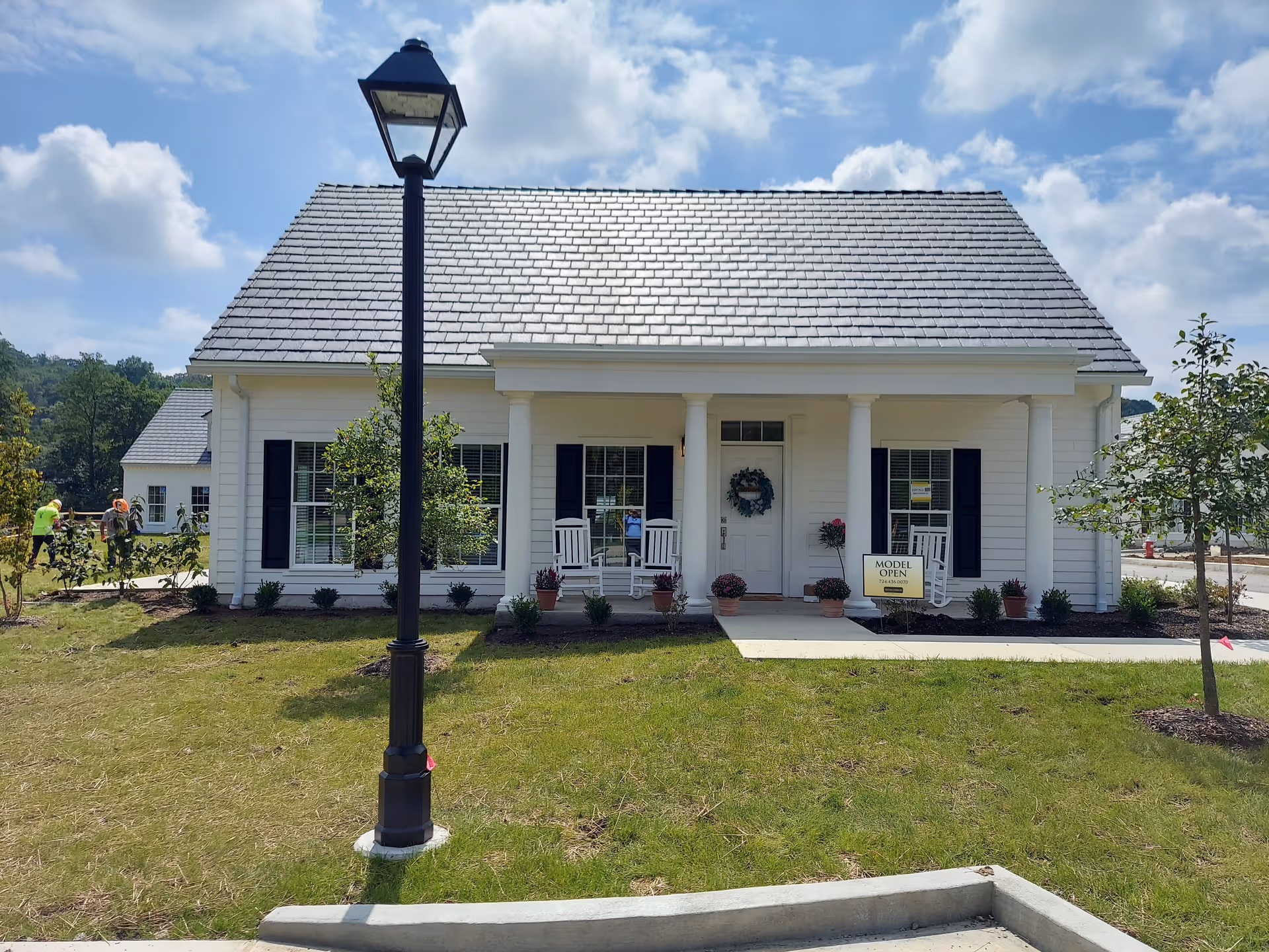 Front exterior view of a white single-story house with a gray shingled roof, black shutters, and a small porch with white rocking chairs and potted plants. A black street lamp stands in the grassy front yard, and a sign near the porch reads 'MODEL OPEN'. Two workers are visible in the background near another building under a partly cloudy sky.