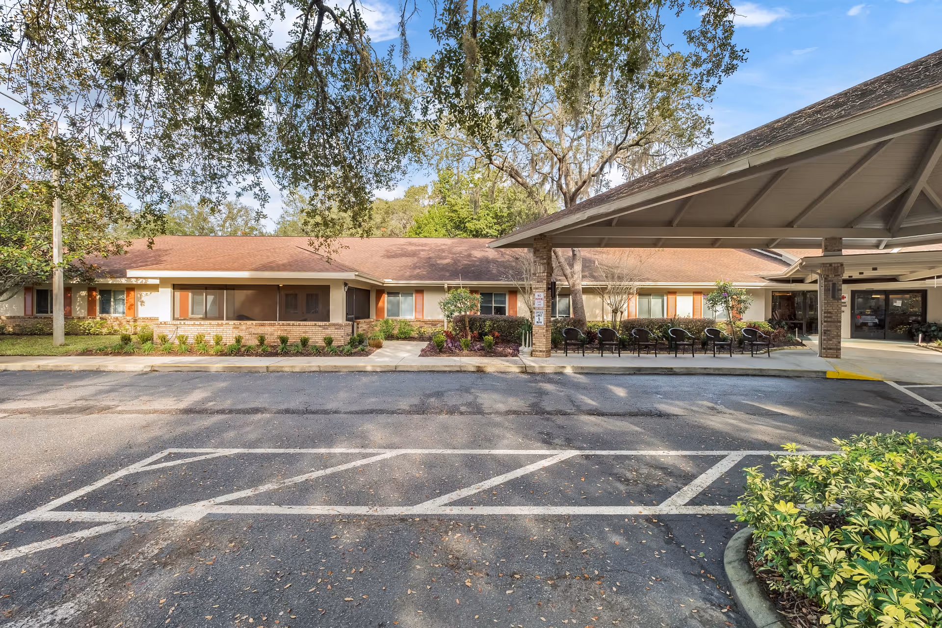 Exterior view of Elevated Estates Forest Oaks facility showing a single-story building with a brown roof and light-colored walls. There is a covered entrance area with several chairs lined up outside. Trees and shrubs surround the building, and a parking area with marked spaces is visible in the foreground under a clear blue sky.
