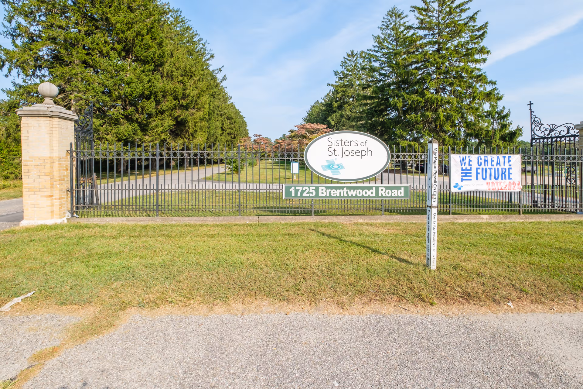 Entrance gate with black iron fencing and brick pillars on either side, a sign reading 'Sisters of St. Joseph' and '1725 Brentwood Road', a banner saying 'We Create The Future Vote 2024', and a white pole with text 'May Peace Prevail On Earth' in multiple languages. Tall green trees and a clear blue sky are visible in the background.