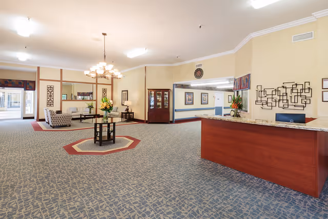 Spacious lobby area of a senior living facility with a reception desk on the right, a round table with a flower arrangement in the center, and a seating area with chairs and a sofa in the background. The room has patterned carpet, beige walls, and decorative wall art.