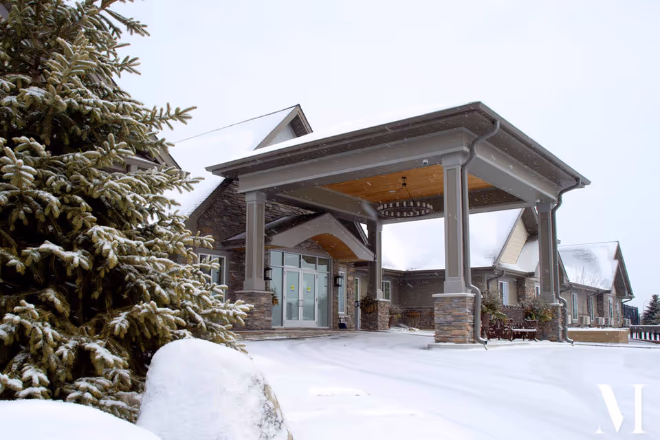 Snow-covered entrance of a building with stone pillars and a covered porch, surrounded by snow-covered trees and ground under a cloudy sky.