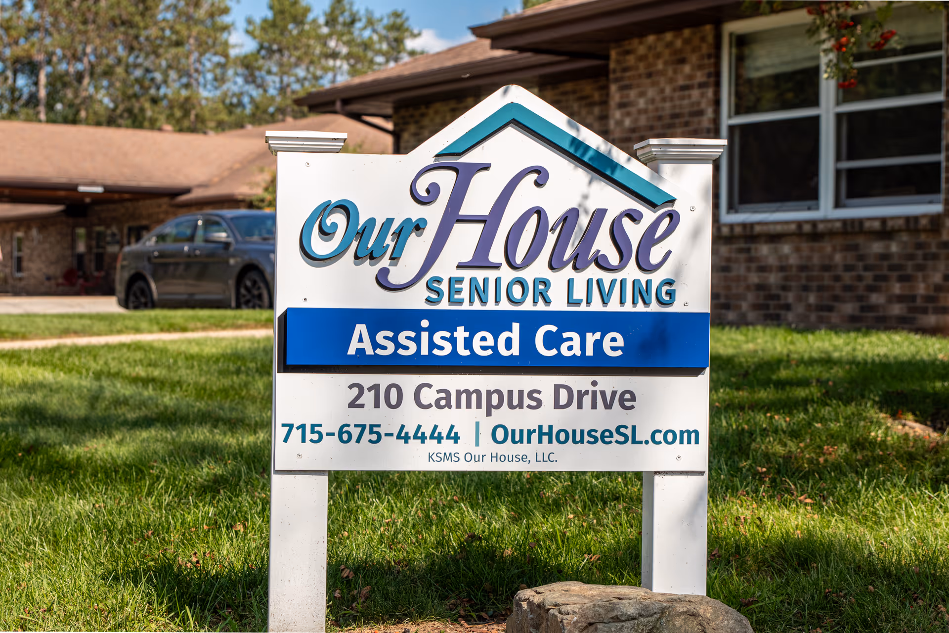 A white sign with blue and teal accents in front of a brick building and green lawn. The sign reads 'Our House Senior Living Assisted Care, 210 Campus Drive, 715-675-4444 | OurHouseSL.com'. A gray car is parked in the background.