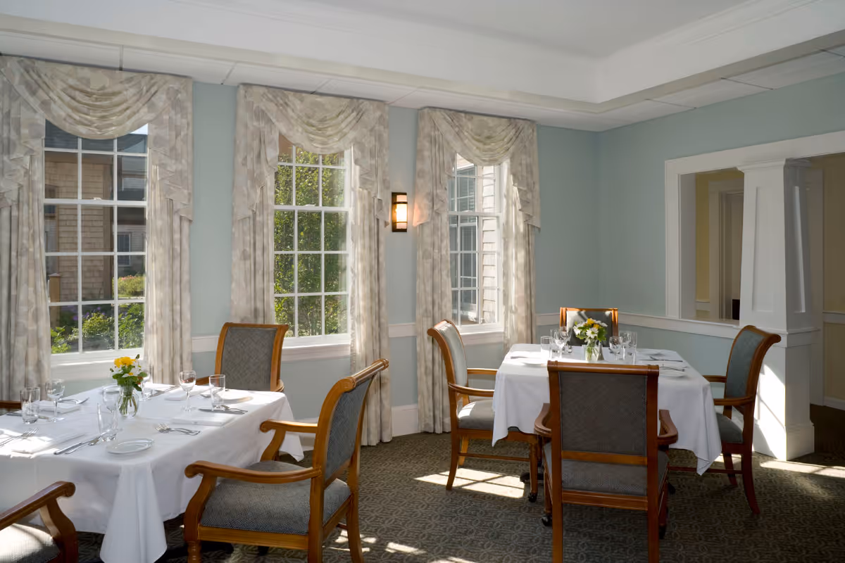 A bright dining room with two tables covered in white tablecloths, each set with glassware, plates, and silverware. Four wooden chairs with cushioned seats surround each table. Three large windows with patterned curtains allow natural light to fill the room. The walls are painted light blue with white trim, and there is a decorative column and wall sconce visible.