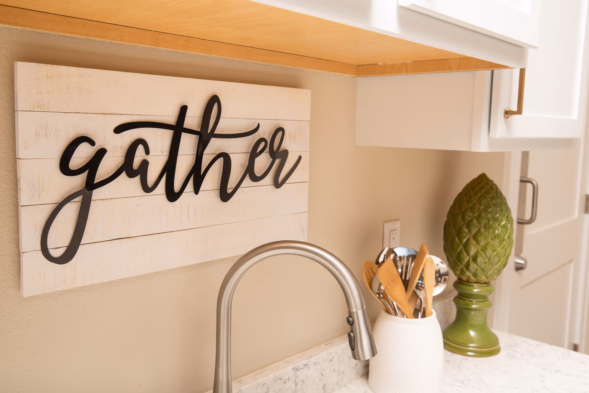 A kitchen countertop with a silver faucet, a white utensil holder containing wooden and metal kitchen utensils, a green decorative artichoke sculpture, and a wall decoration with the word 'gather' in black cursive letters on a white wooden background.